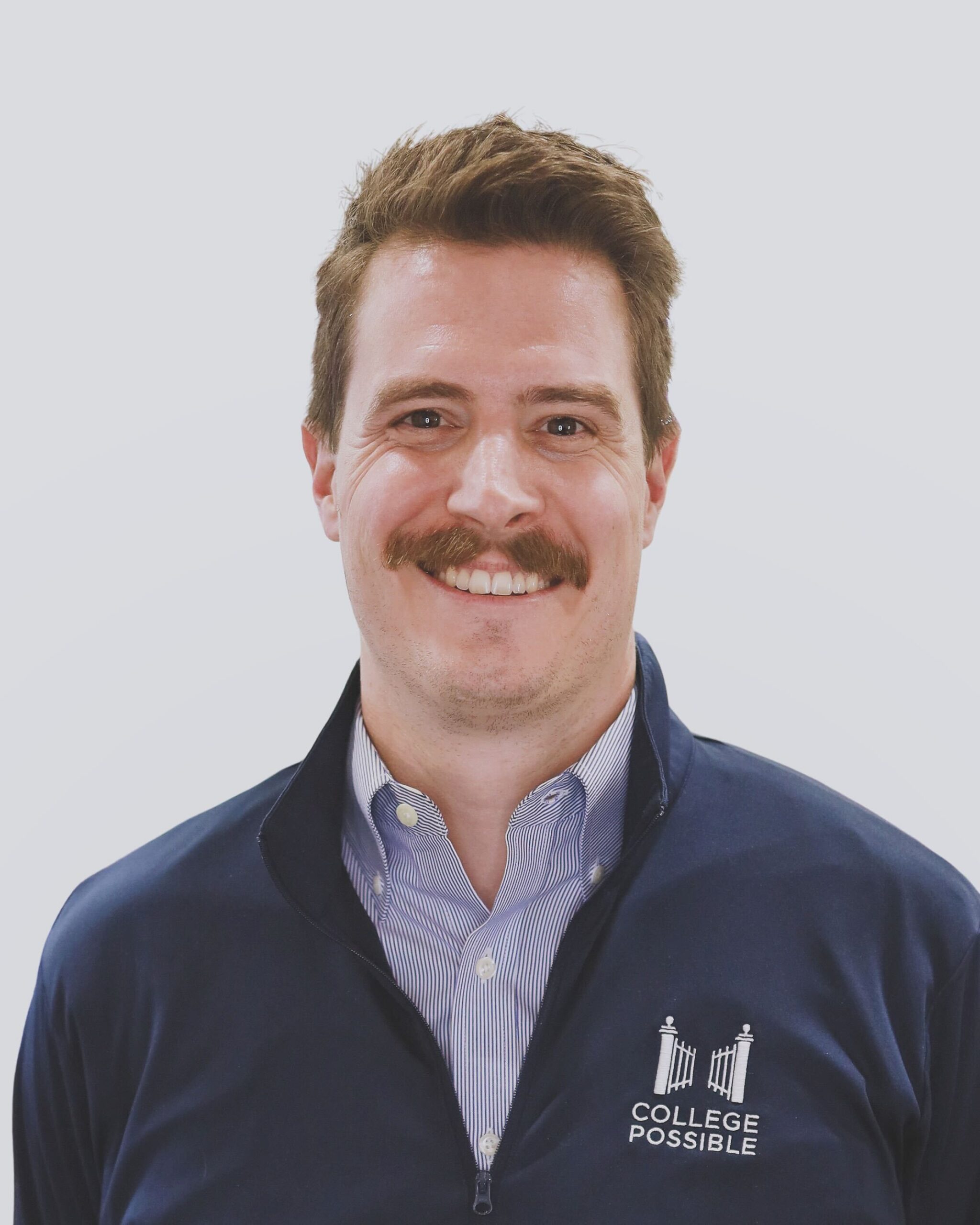 Man in light blue collared shirt and dark blue College Possible branded cardigan stands in front of a white background