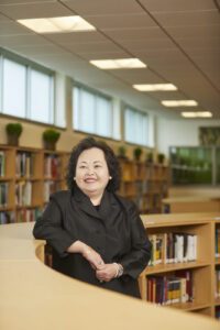 A woman with black hair wearing a black blazer leans on a bookshelf in a library.