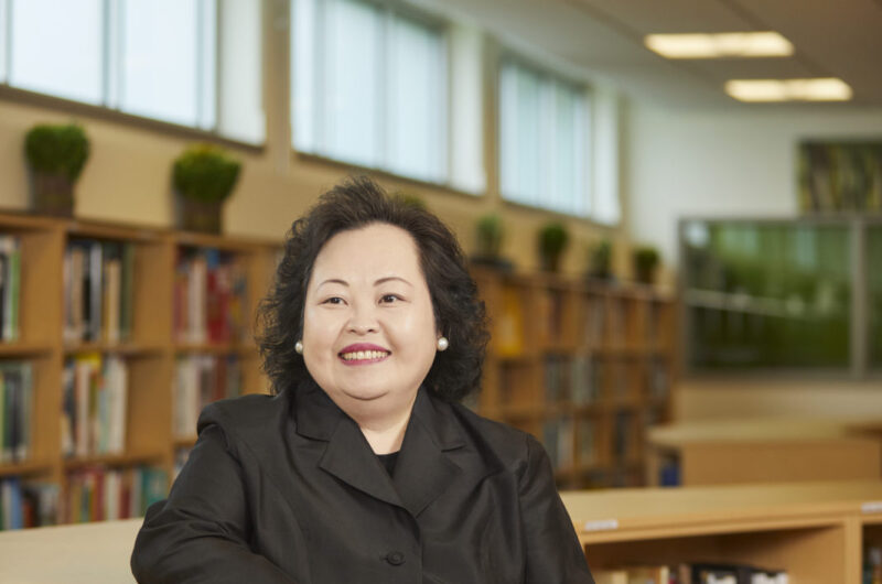 A Woman With Black Hair Wearing A Black Blazer Leans On A Bookshelf In A Library.