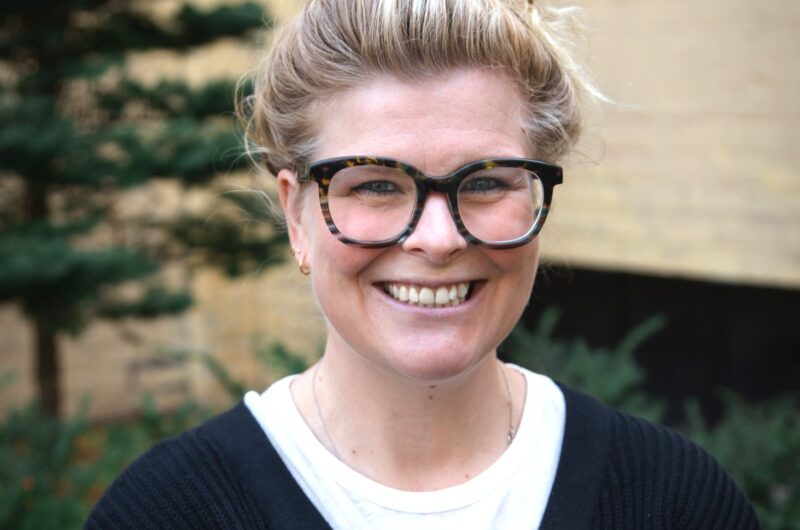 Person With A Bun Hairstyle And Glasses Smiling In Front Of A Brick Building, Wearing A Black Cardigan Over A White Top.