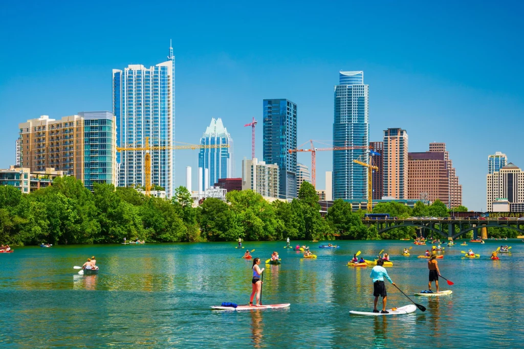 Stand-up paddle boarders float along the Austin skyline on a bright and sunny day. The water is a teal blue and lush green trees line the river walk.