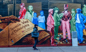 Man walks past colorful mural located in Seventh Ward of Philadelphia. His all-black outfit in contrast to the bright blues, greens and reds of the painted brick.