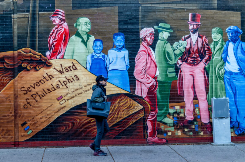 Man Walks Past Colorful Mural Located In Seventh Ward Of Philadelphia. His All-black Outfit In Contrast To The Bright Blues, Greens And Reds Of The Painted Brick.