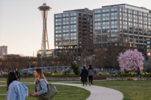 People walk in pairs throughout a park on a walking path. Cherry blossom trees are in bloom in the middle ground. The orange sky highlights the Seattle Space Needle and high-rise building in the background. 