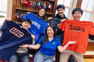 An AmeriCorps coach sits with four high school seniors surrounding her, each holding up a different t-shirt to represent their future college of choice.