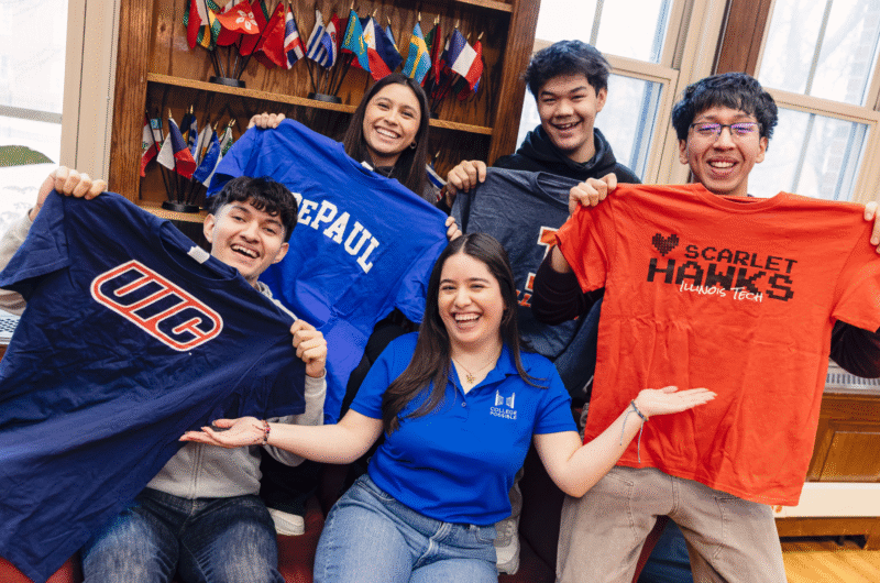 An AmeriCorps coach sits with four high school seniors surrounding her, each holding up a different t-shirt to represent their future college of choice.