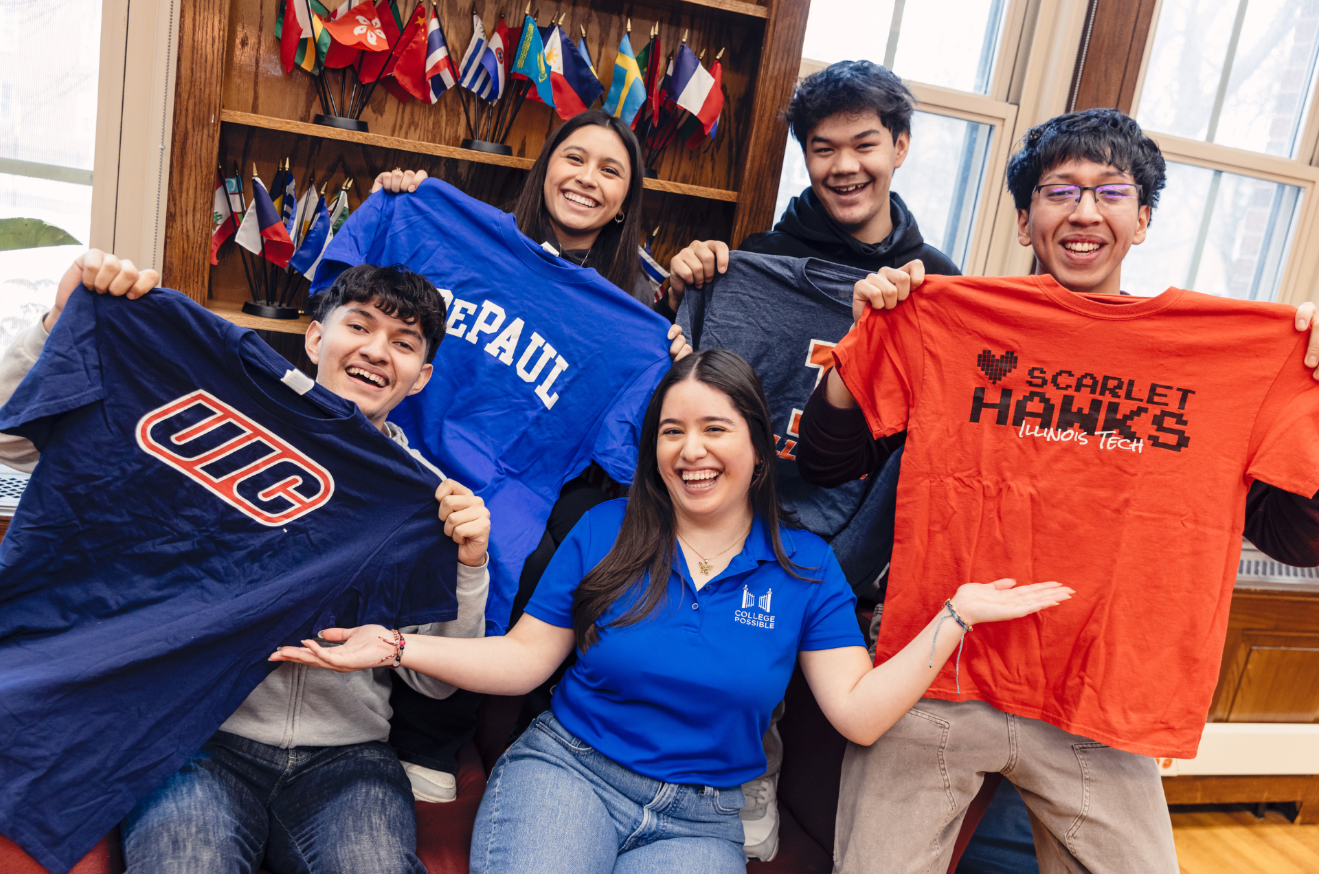 An AmeriCorps coach sits with four high school seniors surrounding her, each holding up a different t-shirt to represent their future college of choice.