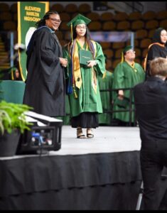 Student in graduation cap and gown on stage holding diploma, shaking the hand of a school official in a robe