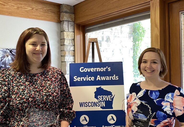 Malysha MacFarland And Lyndsey Johnson Holding AmeriCorps Member Of The Year Awards Standing In Front Of A "Governor's Service Awards" Sign