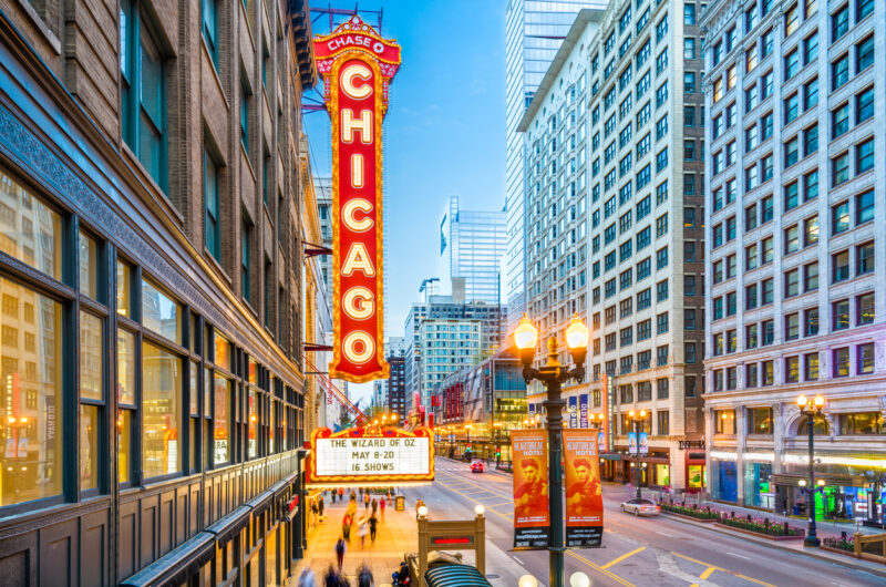 The Landmark Chicago Theatre On State Street Glows, Illuminated By The Twilight Sky. Blurred Pedestrians Line The Sidewalks Next To Chicago’s Skyscrapers.