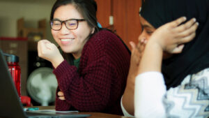 A College Possible AmeriCorps coach and high school student sit at a table together. They both look at a computer screen, laughing with each other. 