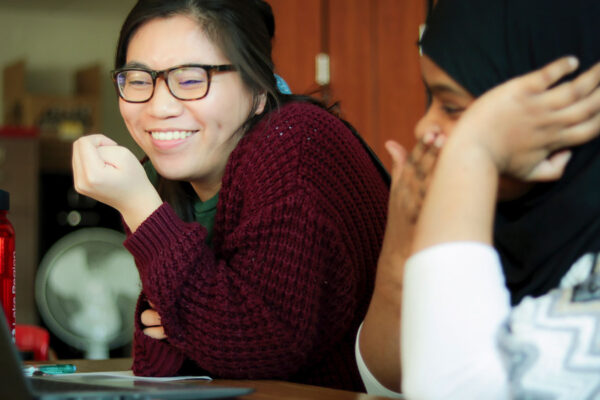 A College Possible AmeriCorps Coach And High School Student Sit At A Table Together. They Both Look At A Computer Screen, Laughing With Each Other.