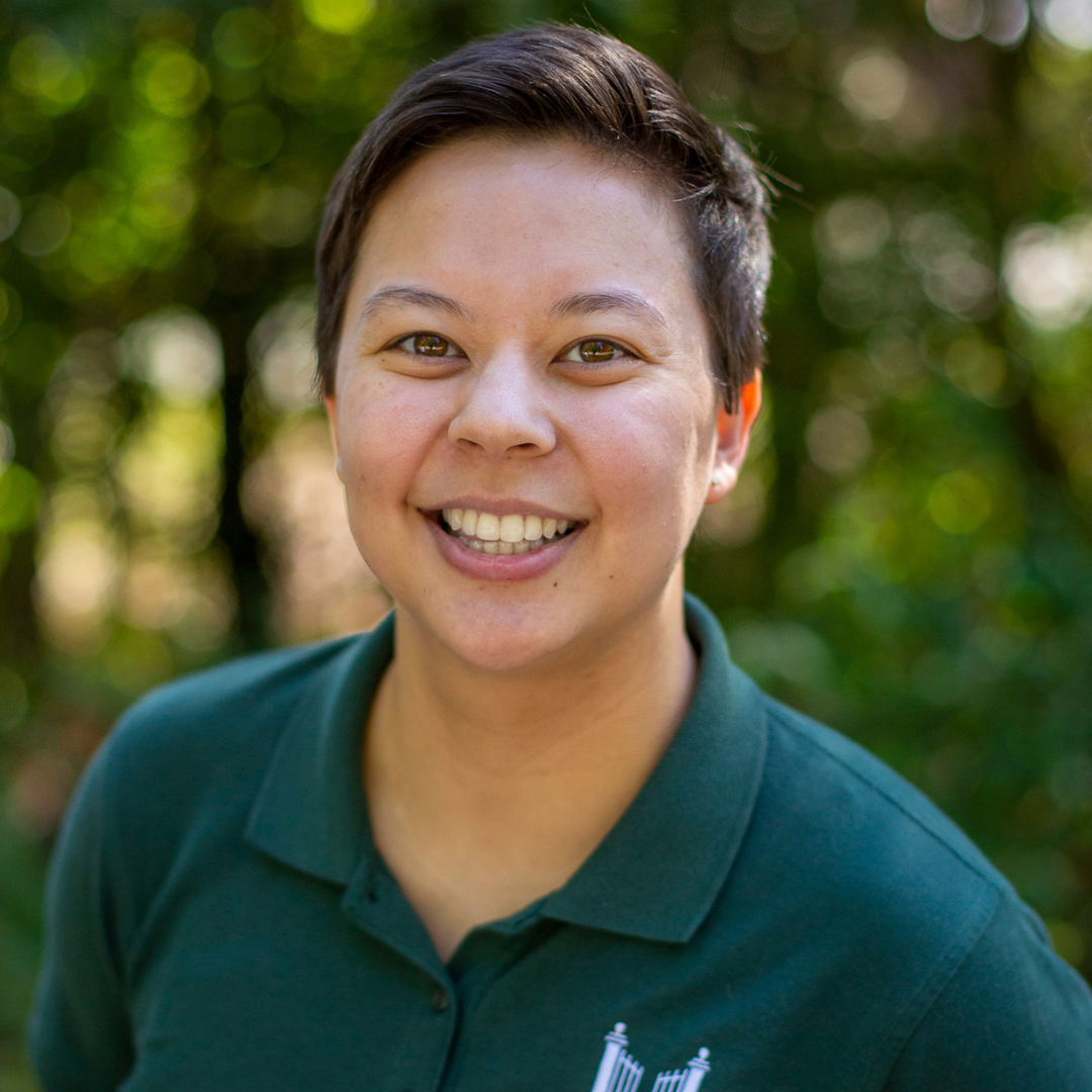 Close up professional headshot of person smiling at camera. They have short brunette hair and are wearing a polo with the College Possible logo on it.
