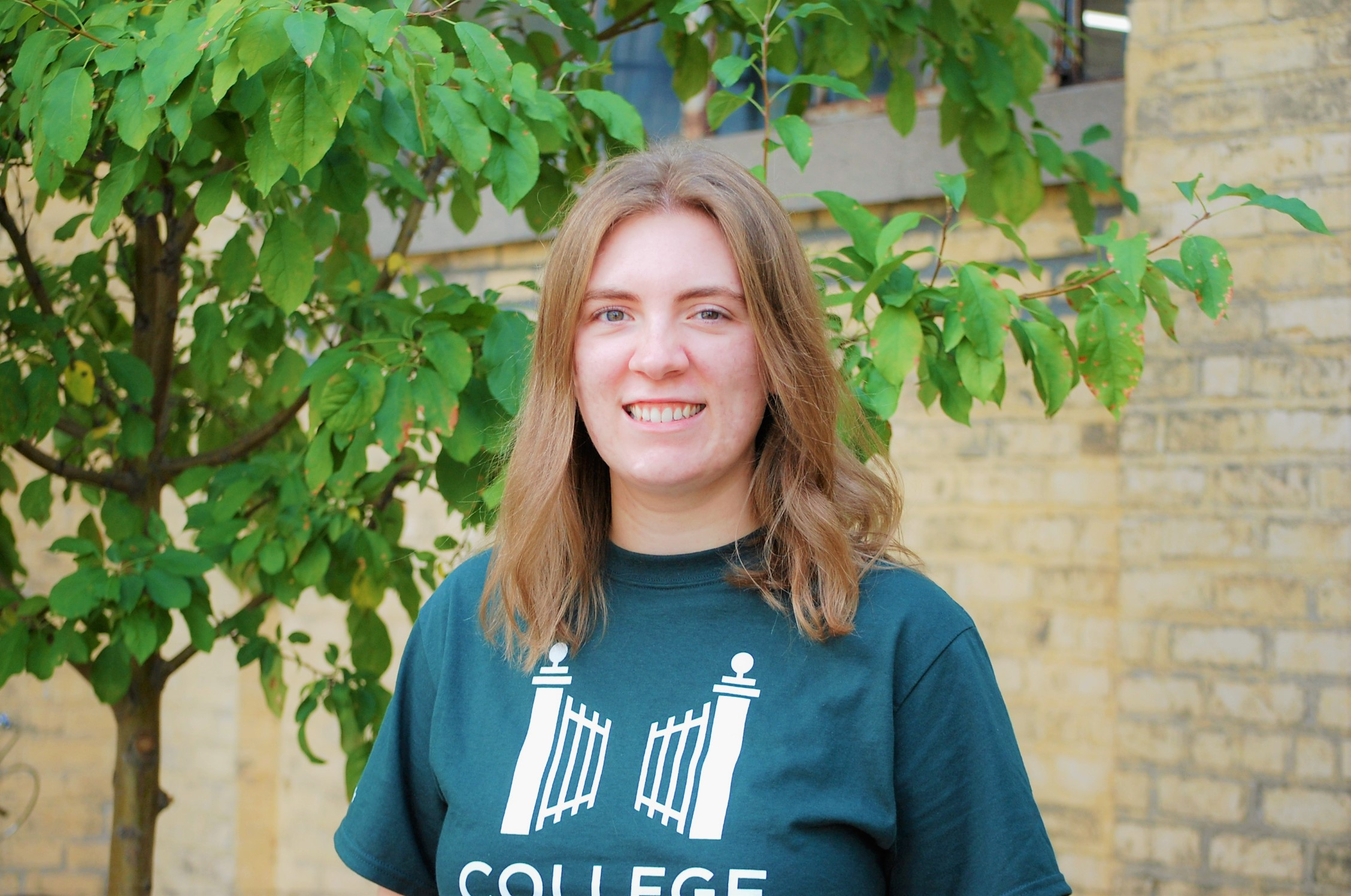Photo of woman in College Possible shirt, with medium length hair that sits at her shoulder. She is standing in front of a tree and posing for a professional headshot.