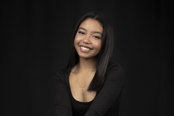 Indoor Photo Of Kyla Posed Sitting Down On A Bench Smiling With A Black Backdrop Behind Her.