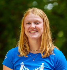 An Image Of A Smiling Woman With Blonde/red Hair, Wearing A Blue T-shirt Standing Outside In Front Of Trees.