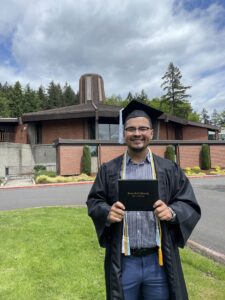 Teo stands in front of a university building wearing a graduation cap and holding a diploma.