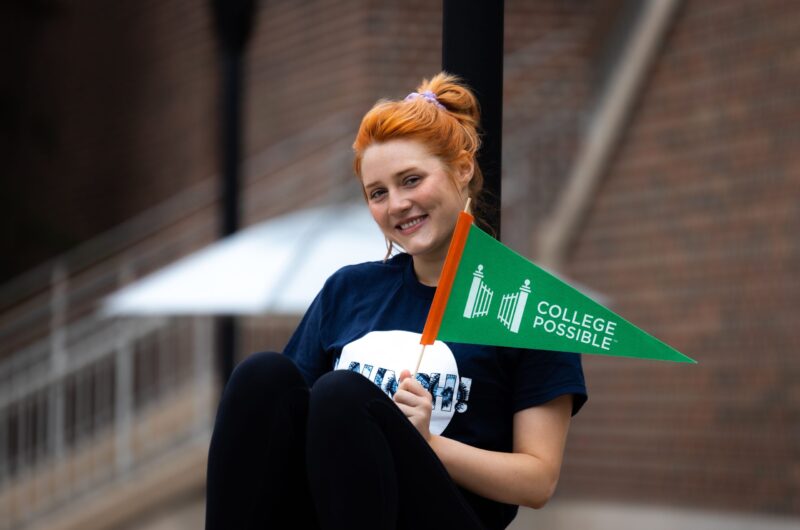College Possible AmeriCorps Coach Sits On A Bench, Smiling While They Hold A College Possible Pennant Flag.