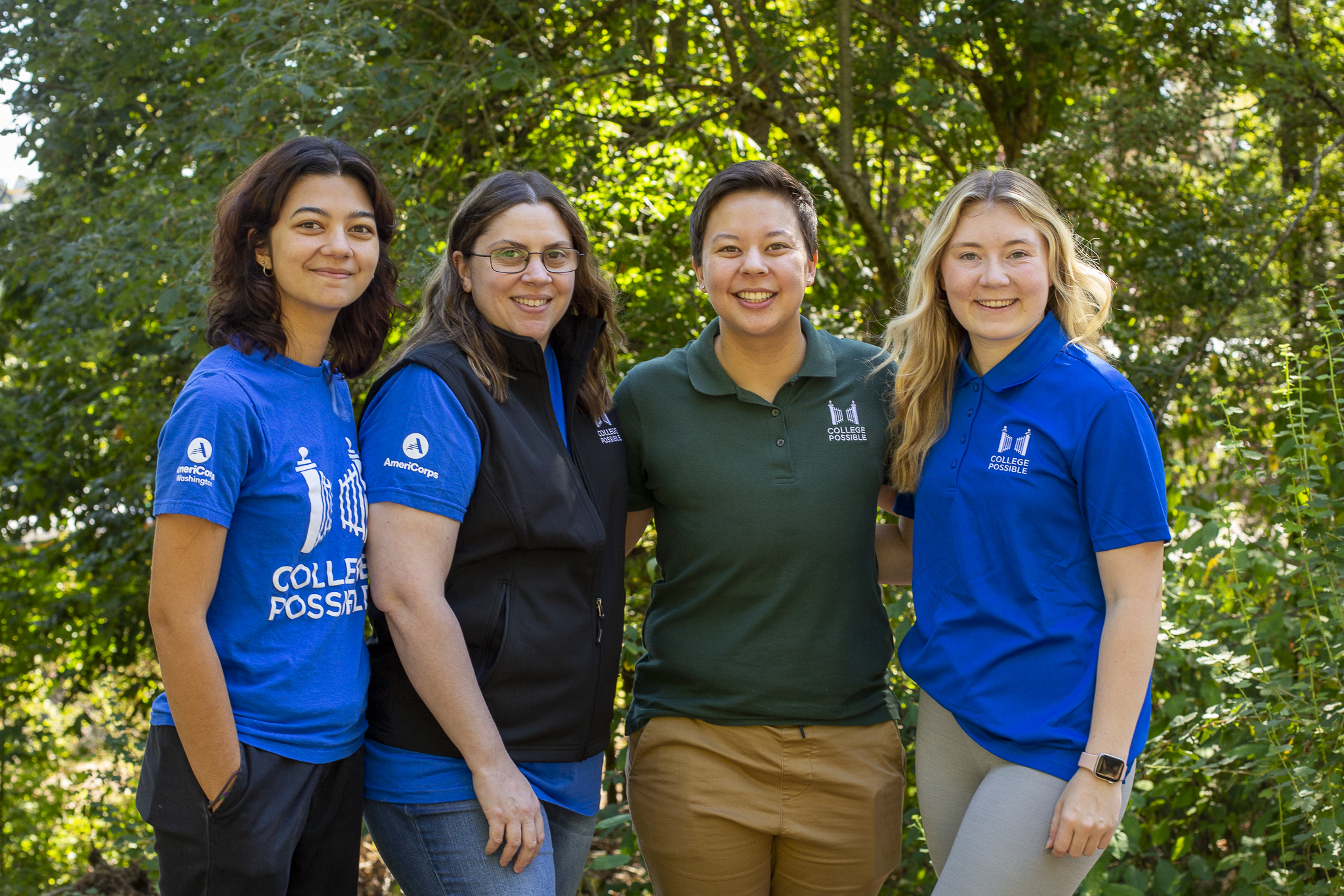 Four women, two with long dark hair, one with short dark hair, and one with long blonde hair, all in College Possible gear, stand smiling at the camera, in front of a green tree filled background.