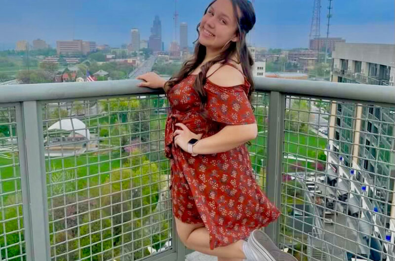 Outdoor Photo Of Gabriella Standing On A Balcony With Cityscape In The Distance.