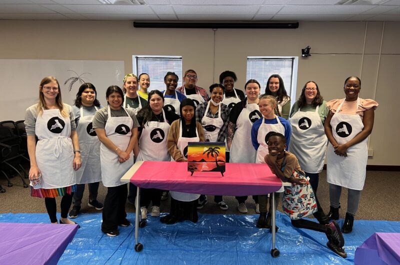 Group Photo Of College Possible Omaha Team In AmeriCorps Aprons Standing Behind A Table With A Painting On It.