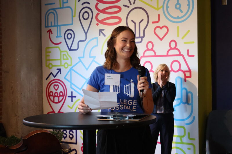 Photo Of Katie Standing At Table/podium Holding A Microphone Smiling At The Audience While Delivering Her Speech.