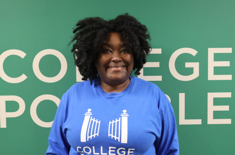 Headshot Photo Of Gwen Wearing Blue College Possible T-shirt With A Green Background Behind Her.