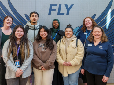8 People Stand In Front Of The Wall Onto Which The Word "fly" Is Written Over An Image Of Wings.