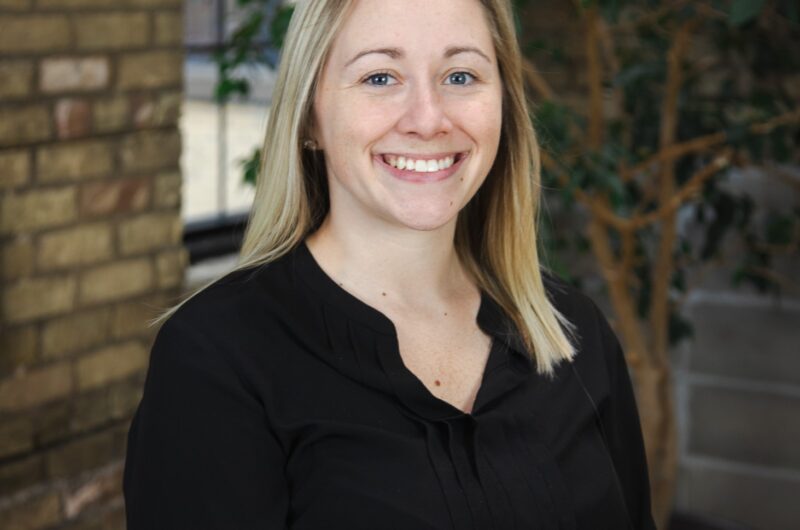 Individual Smiling For A Portrait In Front Of A Brick Wall And Indoor Plants.