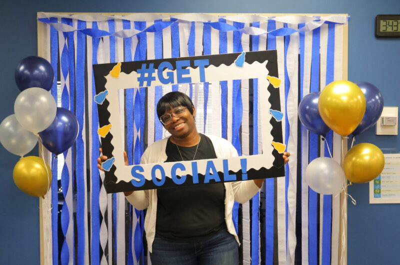 Photo Of T'Netha Holding A Photo Prop Frame With Blue And White Steamers Behind Her And Balloons On Each Side Of Her.