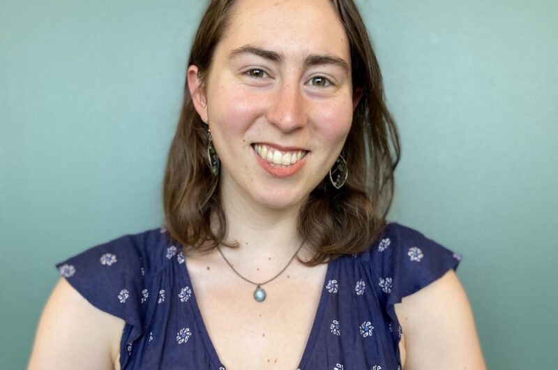 Portrait Of A Smiling Person Wearing A Blue Floral Top Against A Pale Green Background.