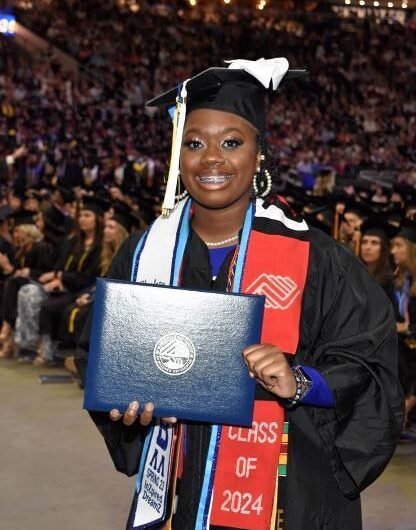 Young Woman Dressed In Graduation Garb, Standing In Front Of A Stadium Full Of People While Smiling And Holding Her Diploma
