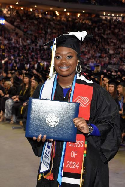 Young woman dressed in graduation garb, standing in front of a stadium full of people while smiling and holding her diploma