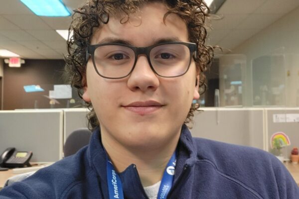 Young Man In His 20s With Curly Brown Hair Wearing AmeriCorps Gear And Smiling At The Camer