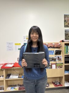 Student wearing a blue sweater stands in a classroom holding a laptop in her hands