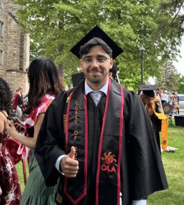 Photo of a graduate on a college campus, giving a thumbs up and wearing a cap and gown with a stole that reads “OLA.”