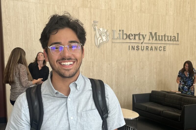 Photo Of A Smiling Person Standing In Front Of A Wall That Reads "Liberty Mutual Insurance"