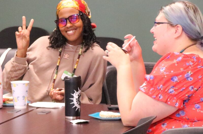 Two People Smiling And Engaging In Conversation At A Table, One Person Making A Peace Sign. They Are Surrounded By Casual Meeting Amenities Like Cups And Snacks.