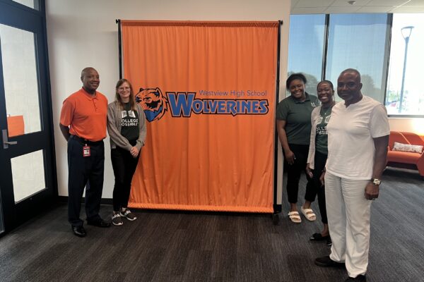 Photo Of College Possible Omaha’s College Access Team Standing With Westview High School Principal In Front Of A Branded Orange Background With The Westview Logo And Mascot.