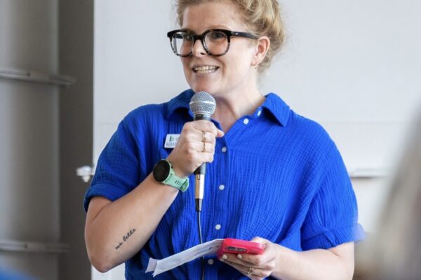 A Person With An Updo Hairstyle Wearing Glasses And A Blue Shirt Speaks Into A Microphone While Holding A Smartphone And Papers, Standing Before An Audience.