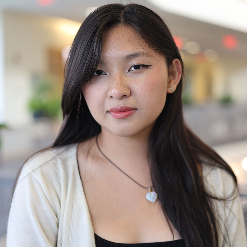 Young person in black shirt and eggshell white cardigan stands in front of an office interior background