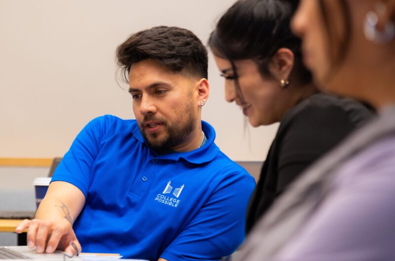 Three People In A Meeting, One Wearing A Blue Shirt, Discussing Content On A Laptop.