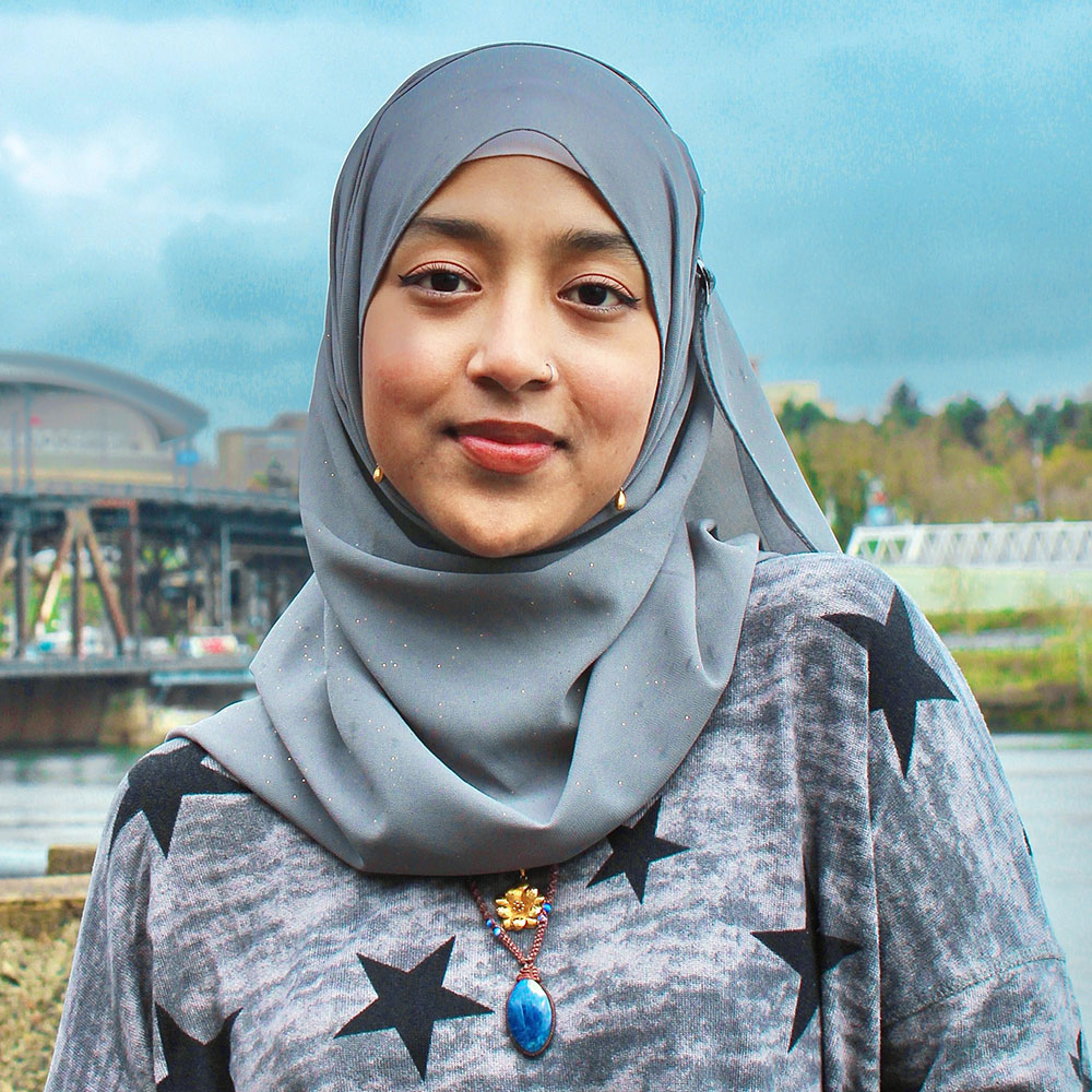 Young woman in a grey headscarf and grey dress with black star pattern stands in front of Portland's inner northeast side waterfront