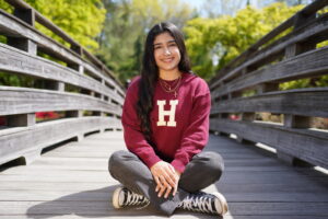 Woman sits cross legged in jeans, tennis shoes and a burgundy Harvard sweatshirt on a wooden footbridge with trees in the background