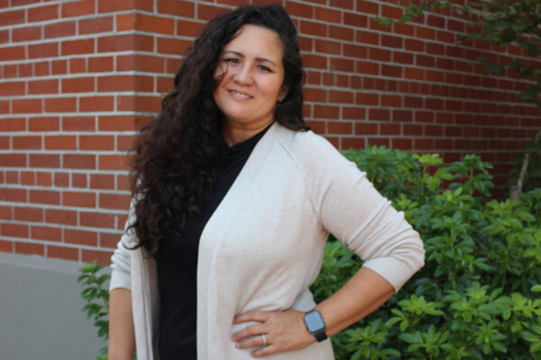 Woman In Black Blouse And White Sweater Stands In Front Of A Brick Schoolhouse Exterior.