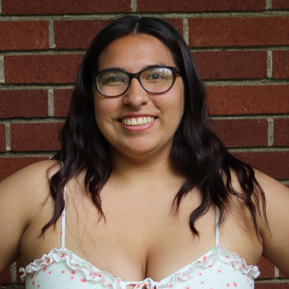 Young woman in white floral camisole dress stands in front of a red brick background