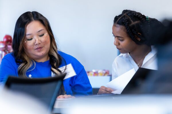 Two Professionals Are Collaborating At An Office Desk, Examining A Document Together. The Setting Appears To Be A Modern Workspace.