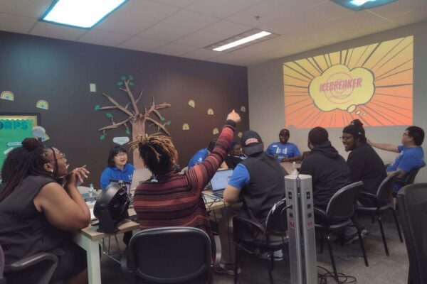 Ten Coaches Gathered Around A Table, Smiling And Chatting While Looking At A Project Screen That Reads “Icebreaker.”