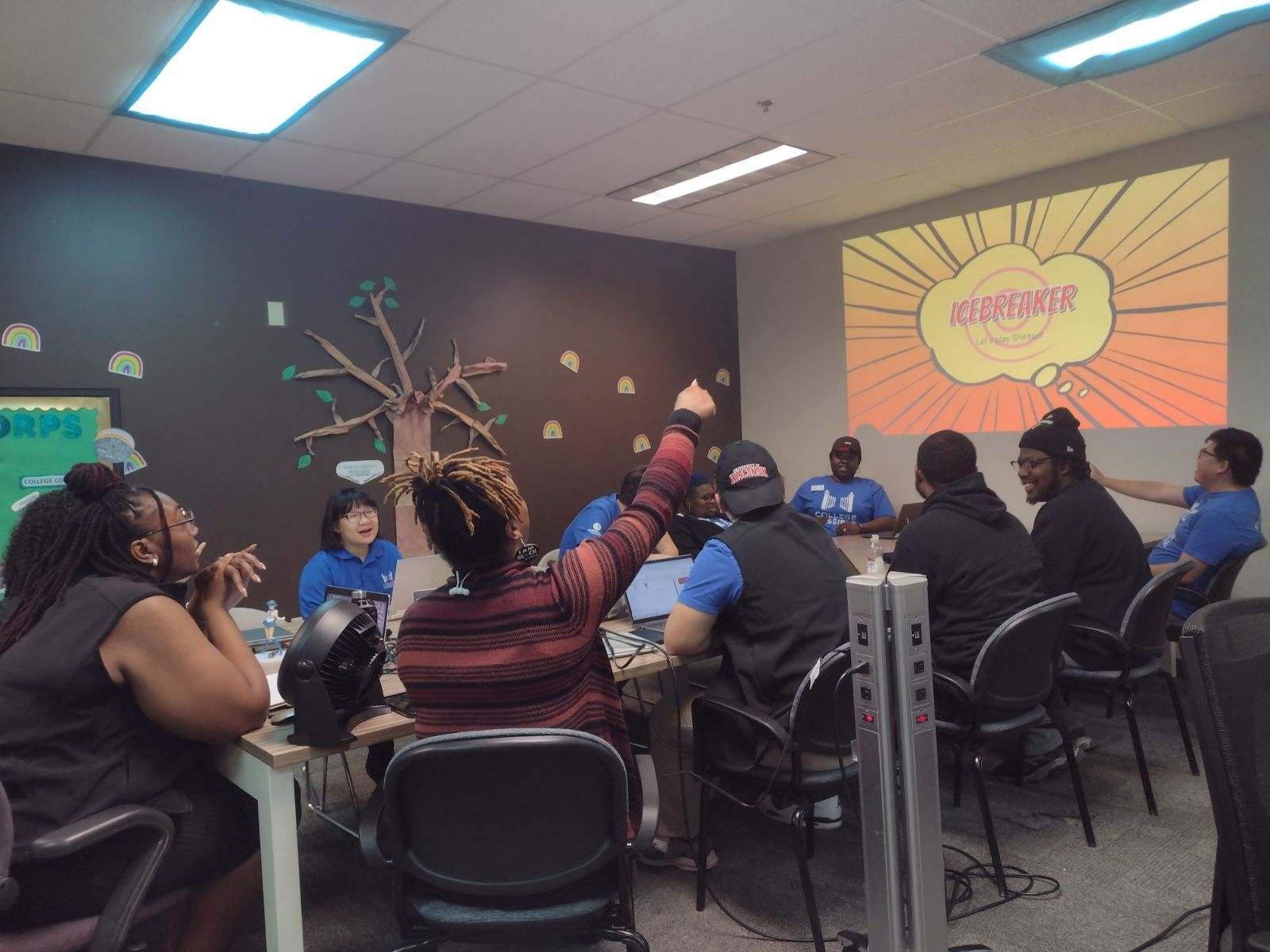 Ten coaches gathered around a table, smiling and chatting while looking at a project screen that reads “Icebreaker.”