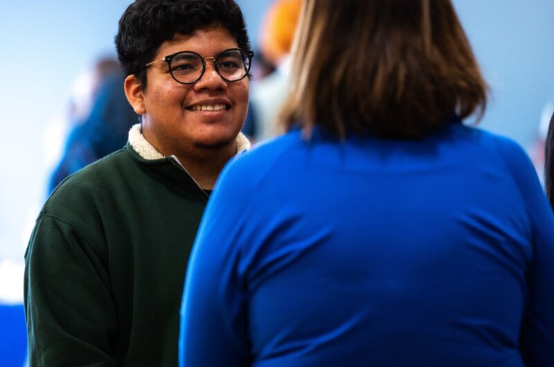 Two People Engaging In A Conversation In A Public Setting, With One Person Wearing Glasses And A Green Sweater Smiling At The Other.